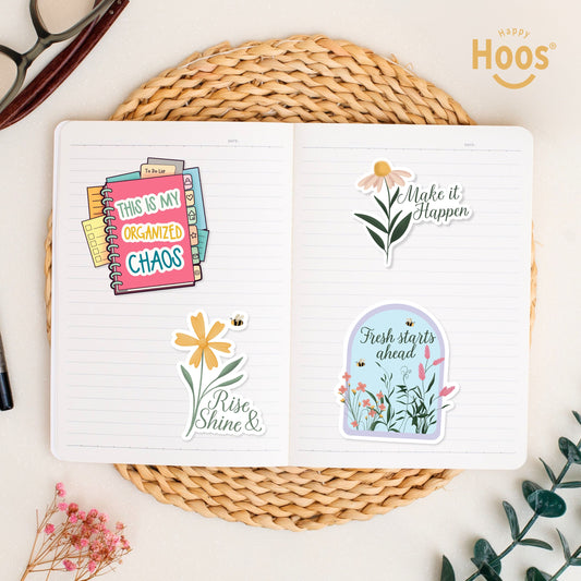 Four stickers in open journal on woven placemat: 'This is My Organized Chaos' planner, 'Rise Shine &' yellow flower, 'Make it Happen' daisy, and 'Fresh starts ahead' floral arch