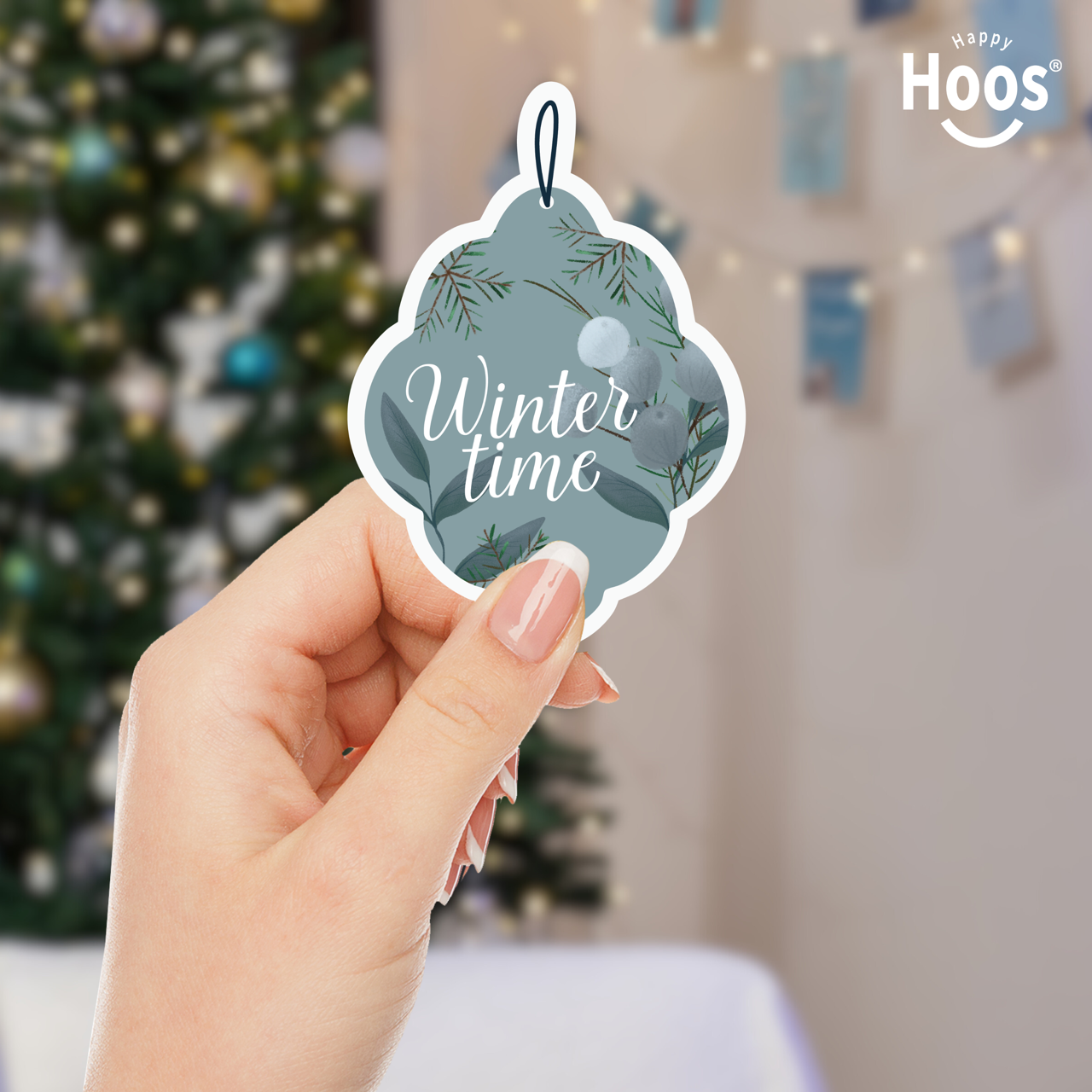 Hand holding the Winter Ornament Sticker with 'Winter time' text in front of a Christmas tree.