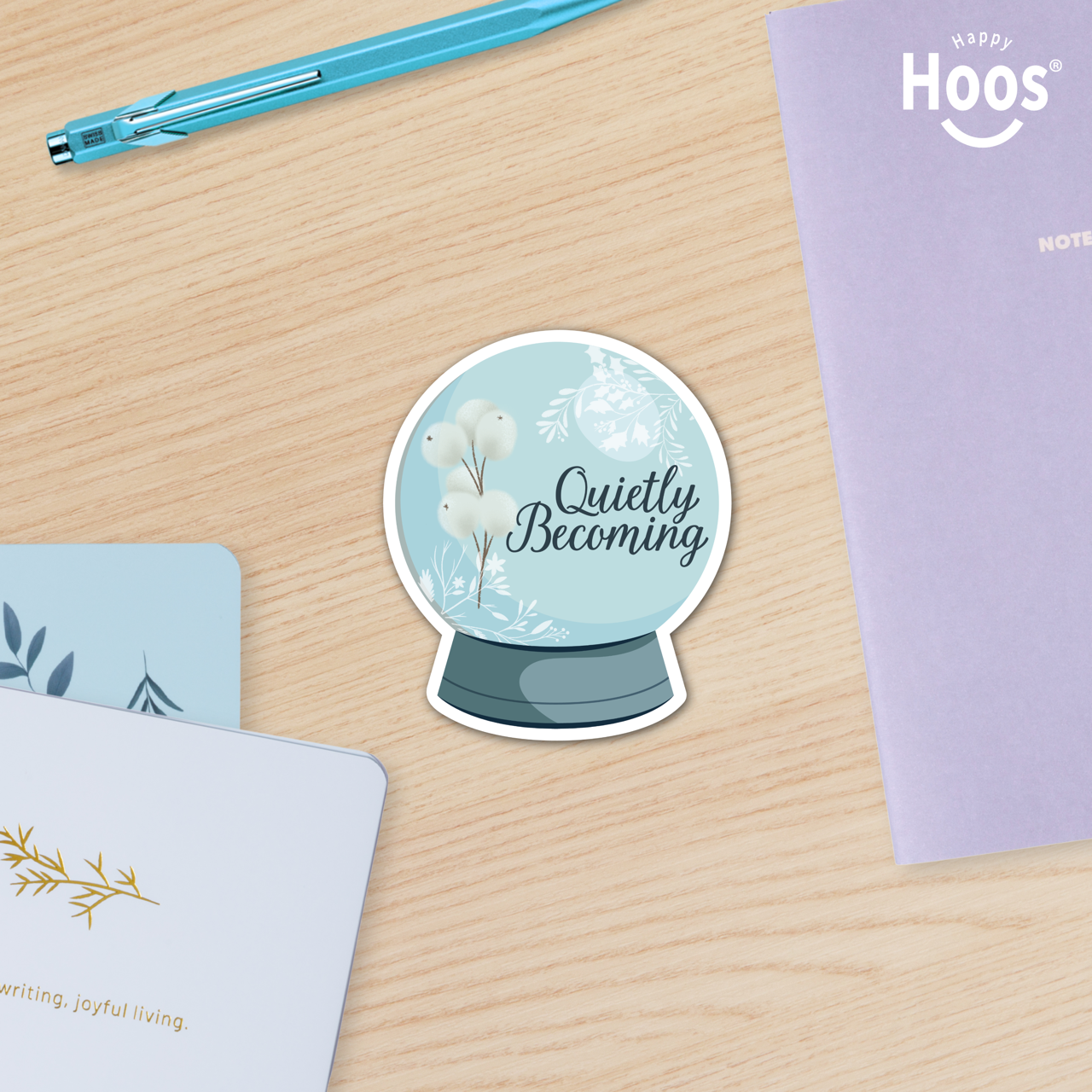 Sticker with a snow globe design and words "Quietly Becoming" and text on a wooden surface with stationery items.