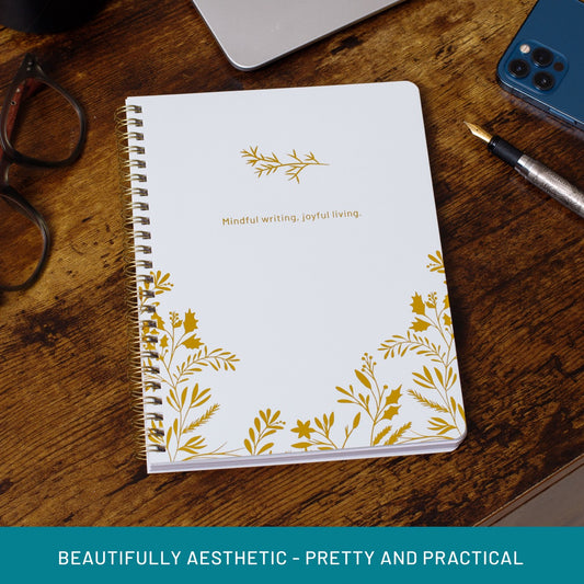 White winter botanical spiral notebook with gold foil and quote ‘Mindful writing, joyful living on a wooden desk with glasses and phone.