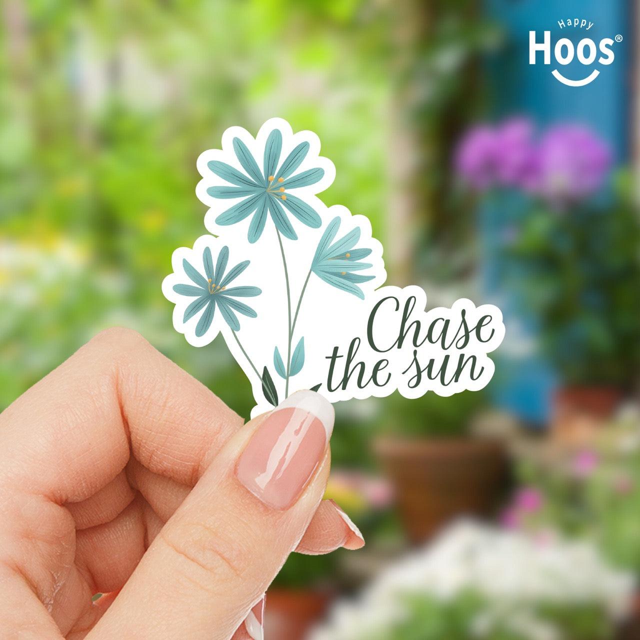 Hand holding a sticker with blue flowers and 'Chase the sun' text against a garden background.