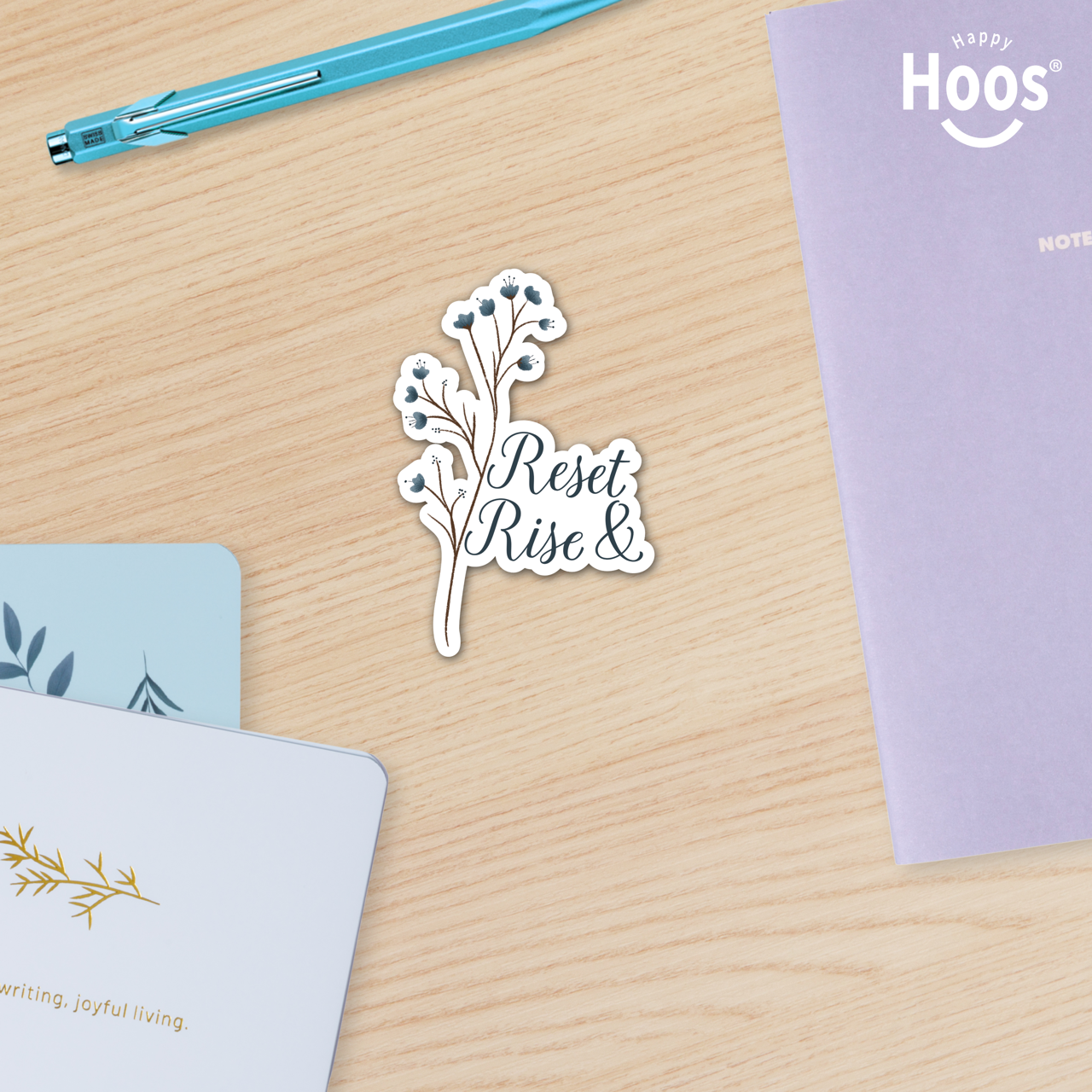 Sticker with 'Reset Rise &' text on a wooden surface with stationery items.