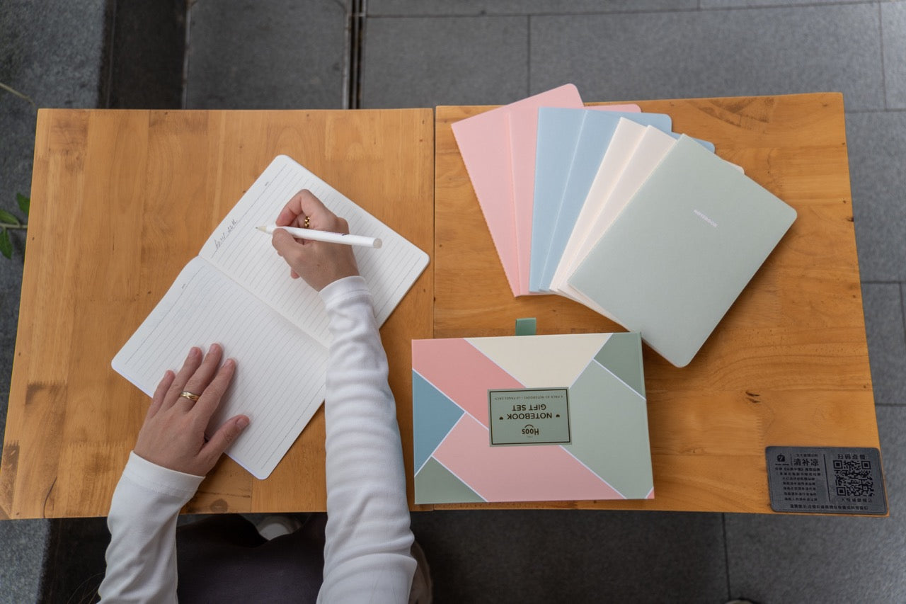 Person writing in a notebook with colorful notebooks and a box on a wooden table.
