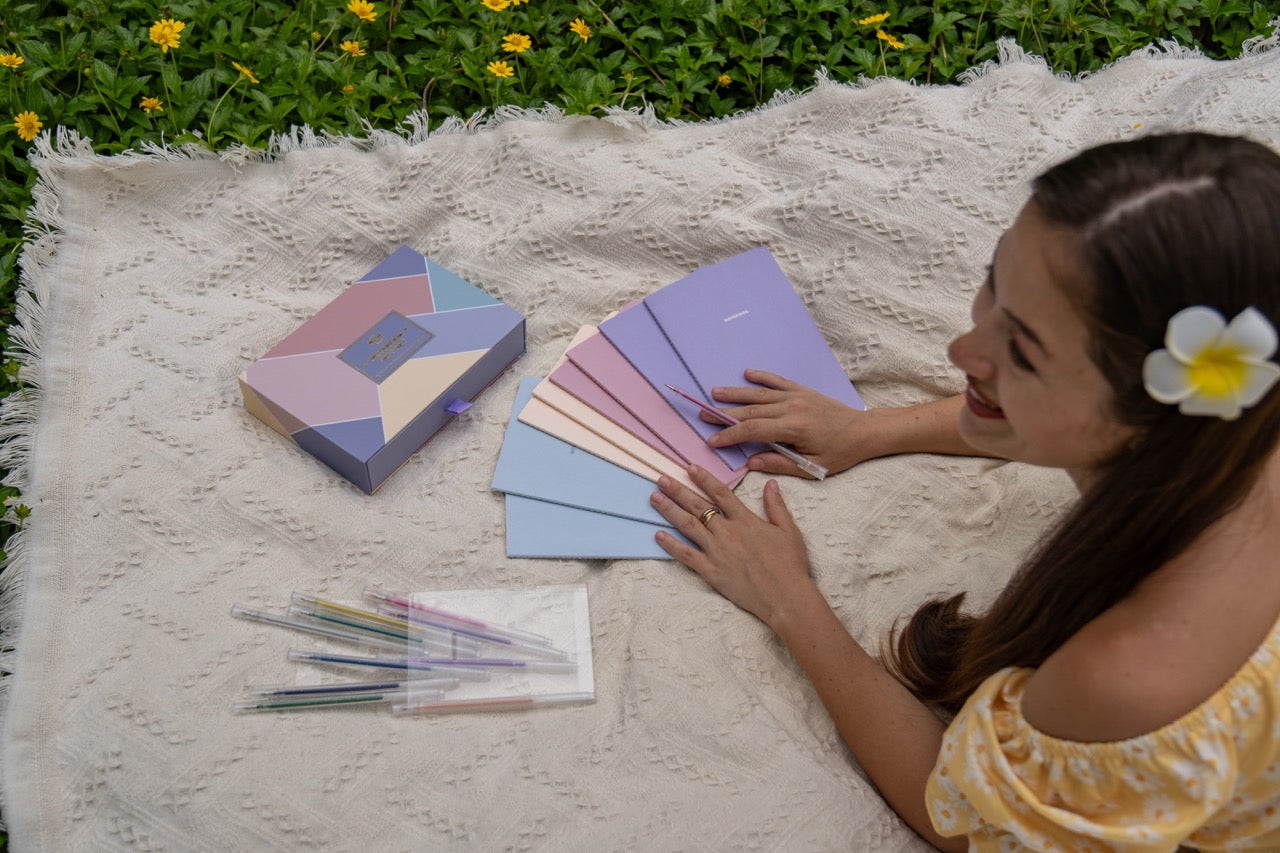 Woman sitting on a blanket with color summer notebooks, a geometric beautiful box  and a box of crayons outdoors.