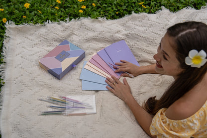 Woman sitting on a blanket with color summer notebooks, a geometric beautiful box  and a box of crayons outdoors.