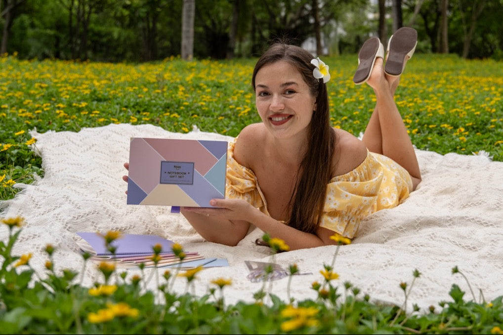 Woman in a yellow dress lying on a white blanket in a grassy field with flowers, holding a product box.