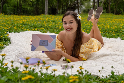 Woman in a yellow dress lying on a white blanket in a grassy field with flowers, holding a product box.