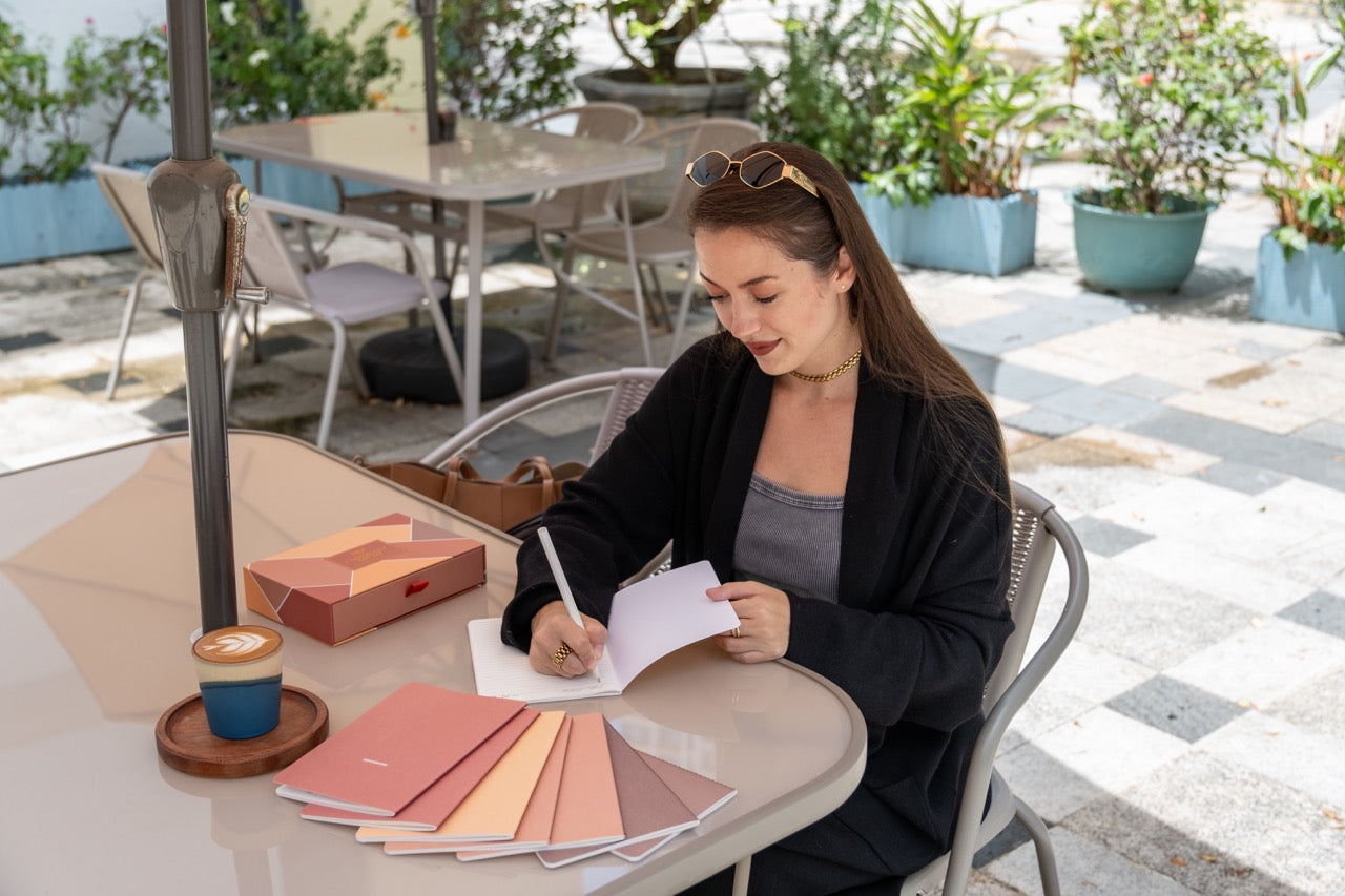 Woman sitting at an outdoor cafe table with 8 fall notebooks, writing on one of them with the box next to her and a cup of coffee.