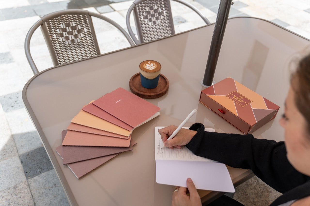 Person writing in a notebook at an outdoor table with coffee and notebooks.
