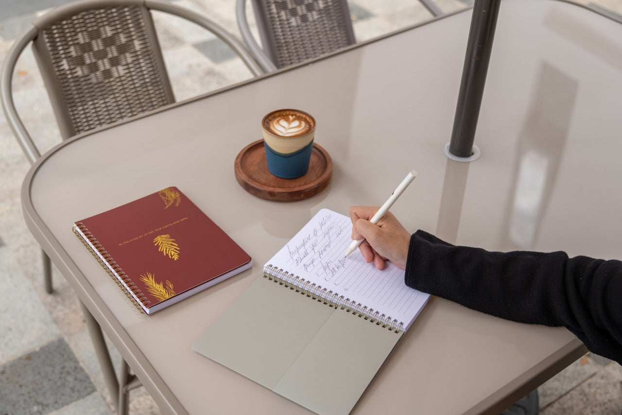 Person writing in a notebook with a coffee cup and another notebook on a table outdoors.
