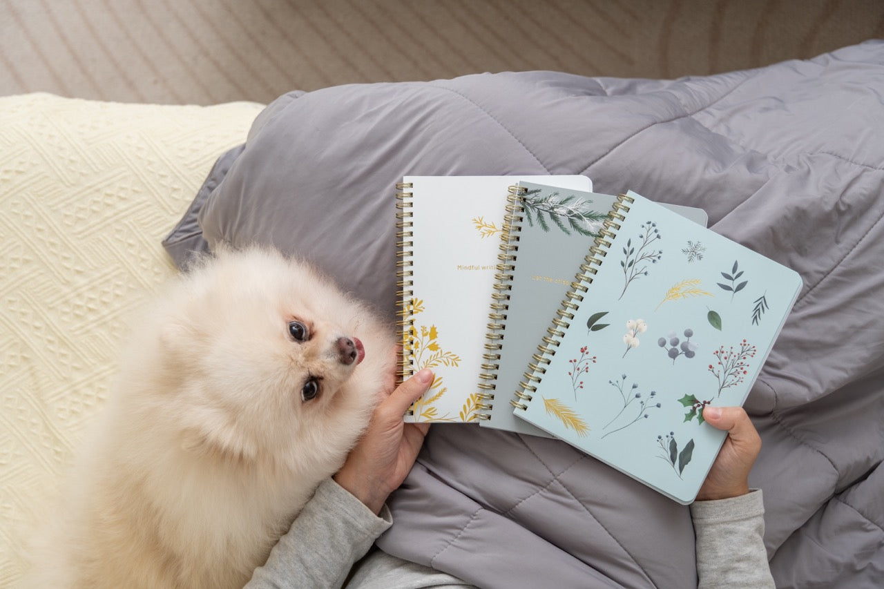 Person holding three spiral-bound notebooks with floral designs on a bed, accompanied by a small white dog.