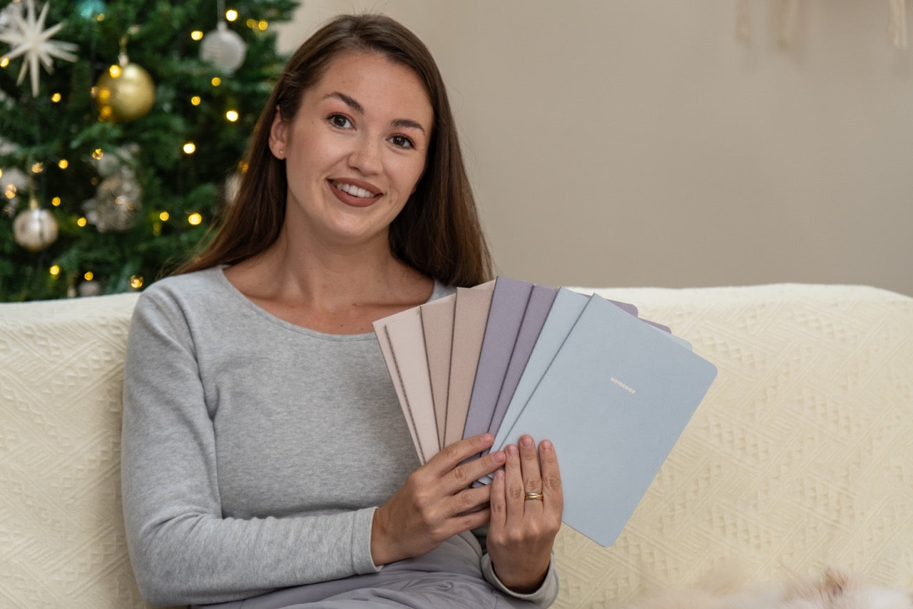 Woman holding 8 winter themed notebooks in front of a decorated Christmas tree