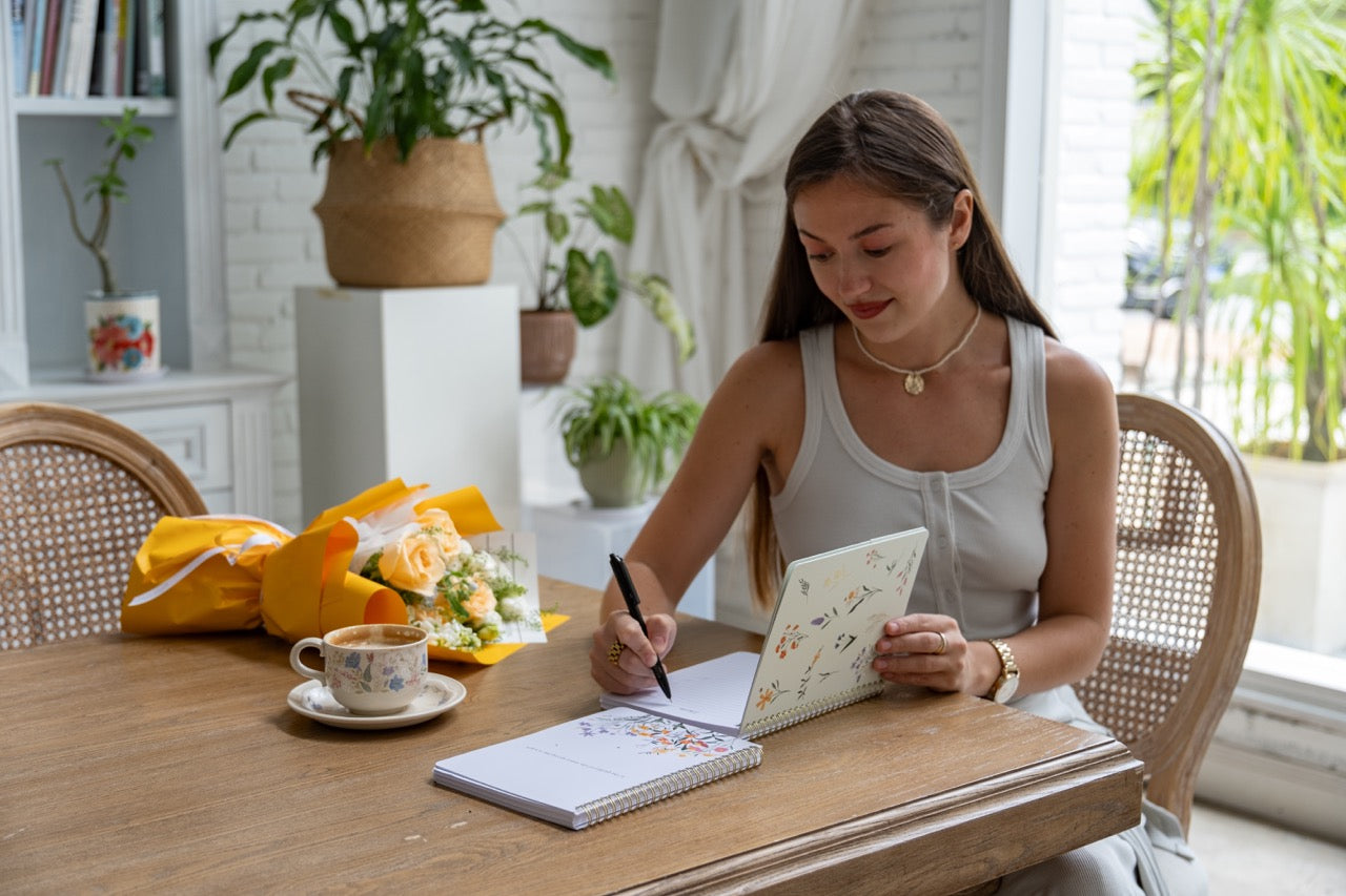 Woman sitting at a table with a notebook and pen, surrounded by plants in a bright room.