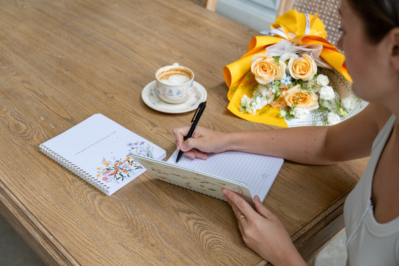 Person writing in a notebook with a cup of coffee and flowers on a wooden table