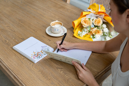 Person writing in a notebook with a cup of coffee and flowers on a wooden table