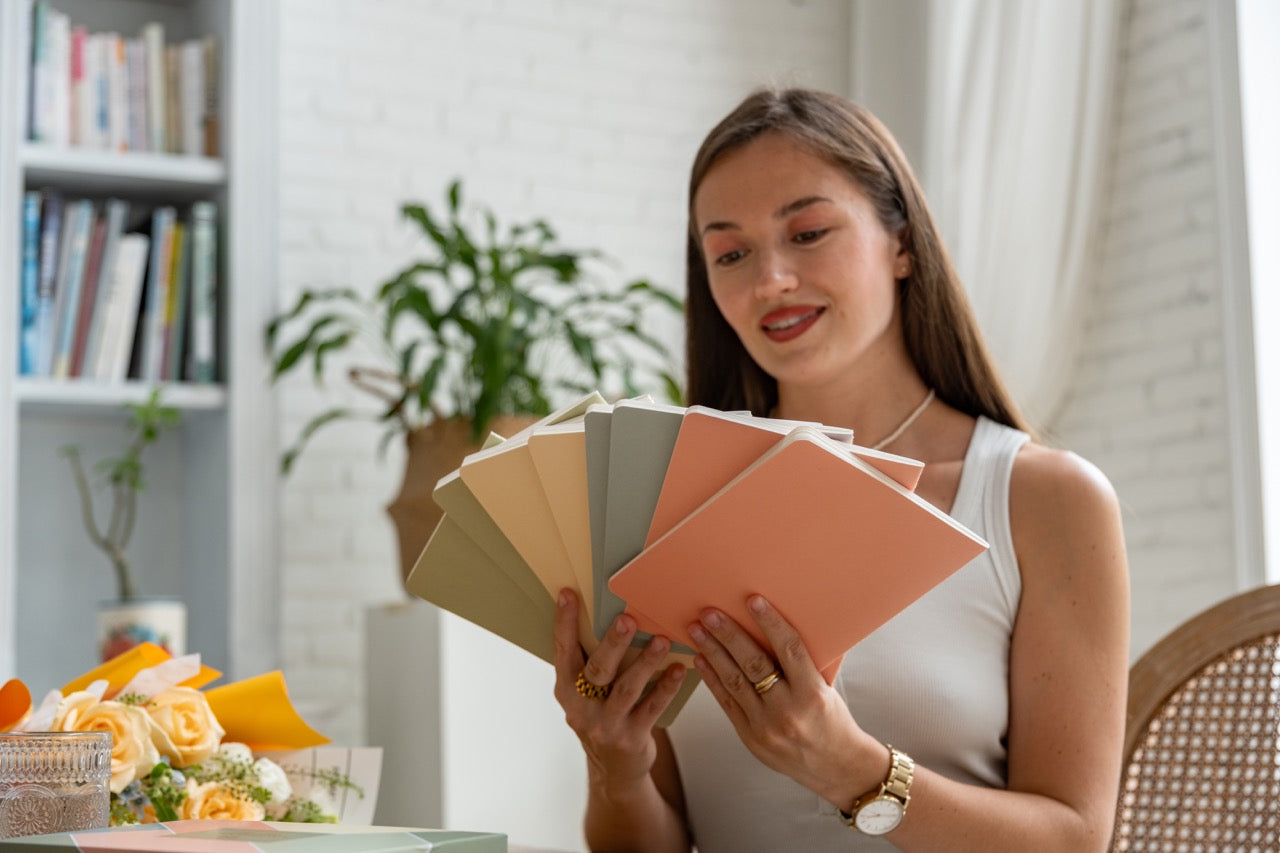 Woman holding 8 spring notebook journals in a bright room with plants and books.