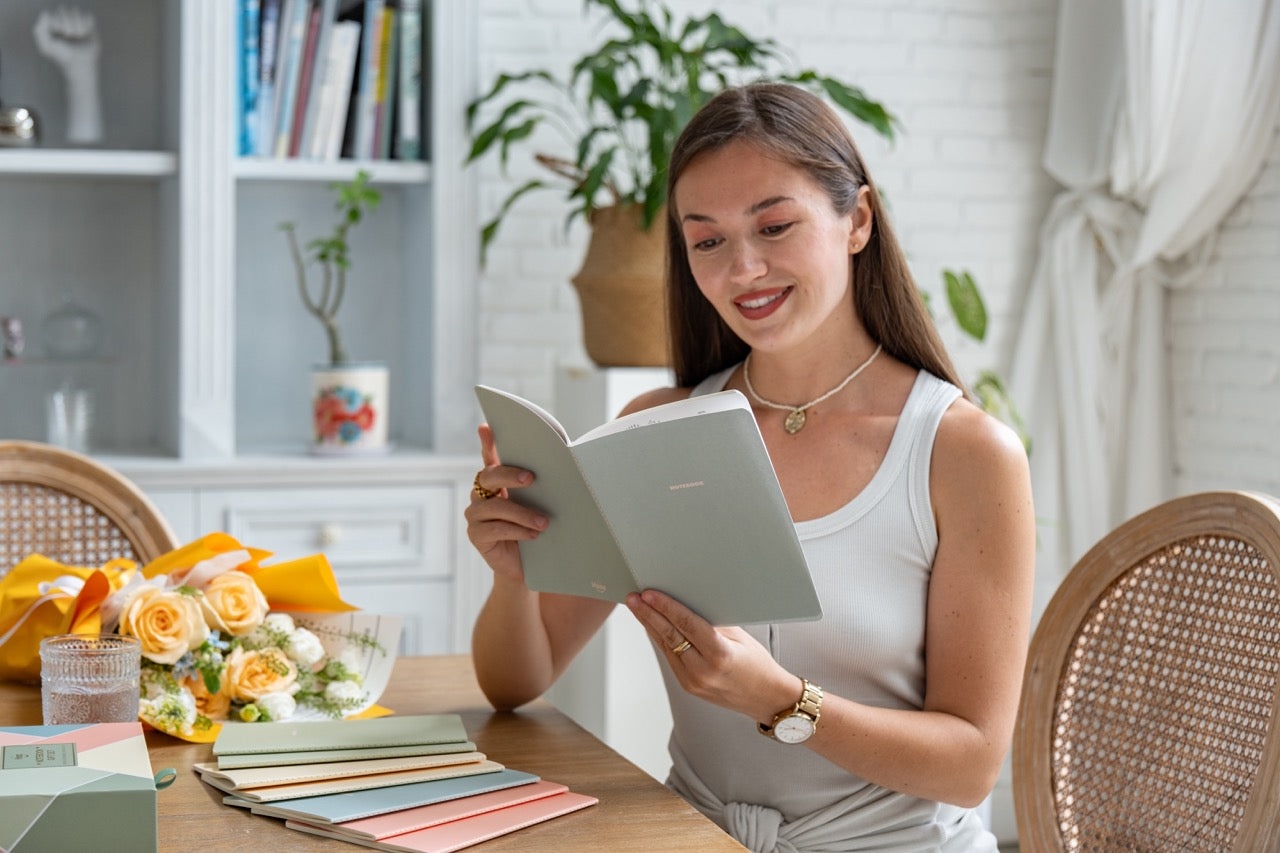 Woman reading a spring colored notebook in a cozy room with a table and on top of the table are the 7 other notebooks and colorful box