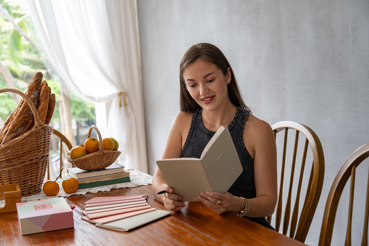Woman reading her notebook at a table with a basket of bread and oranges in a bright room while a beautiful box and 7 other. notebooks are on the table.