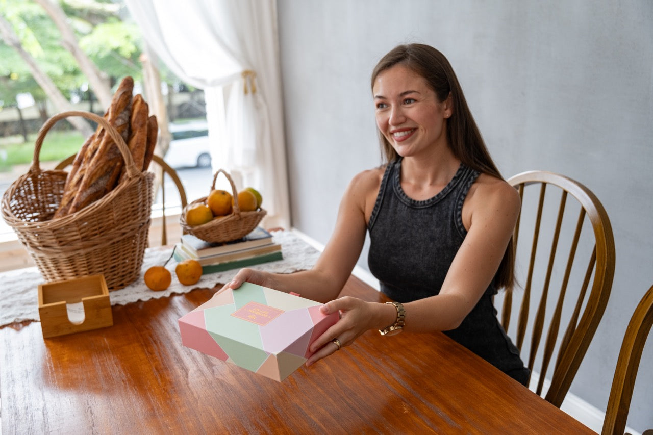 Woman sitting at a table with a box, surrounded by baskets of bread and oranges.