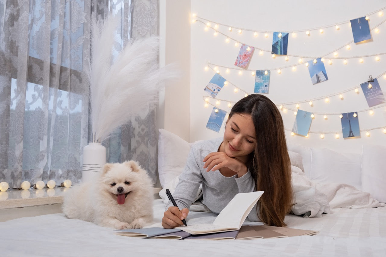 Woman lying on a bed with a dog, surrounded by string lights and decorative items writing on winter-themed notebooks.