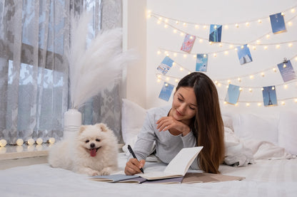 Woman lying on a bed with a dog, surrounded by string lights and decorative items writing on winter-themed notebooks.