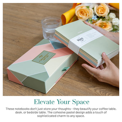 Pastel-colored notebooks with a hand holding one, on a wooden surface with flowers and a glass.