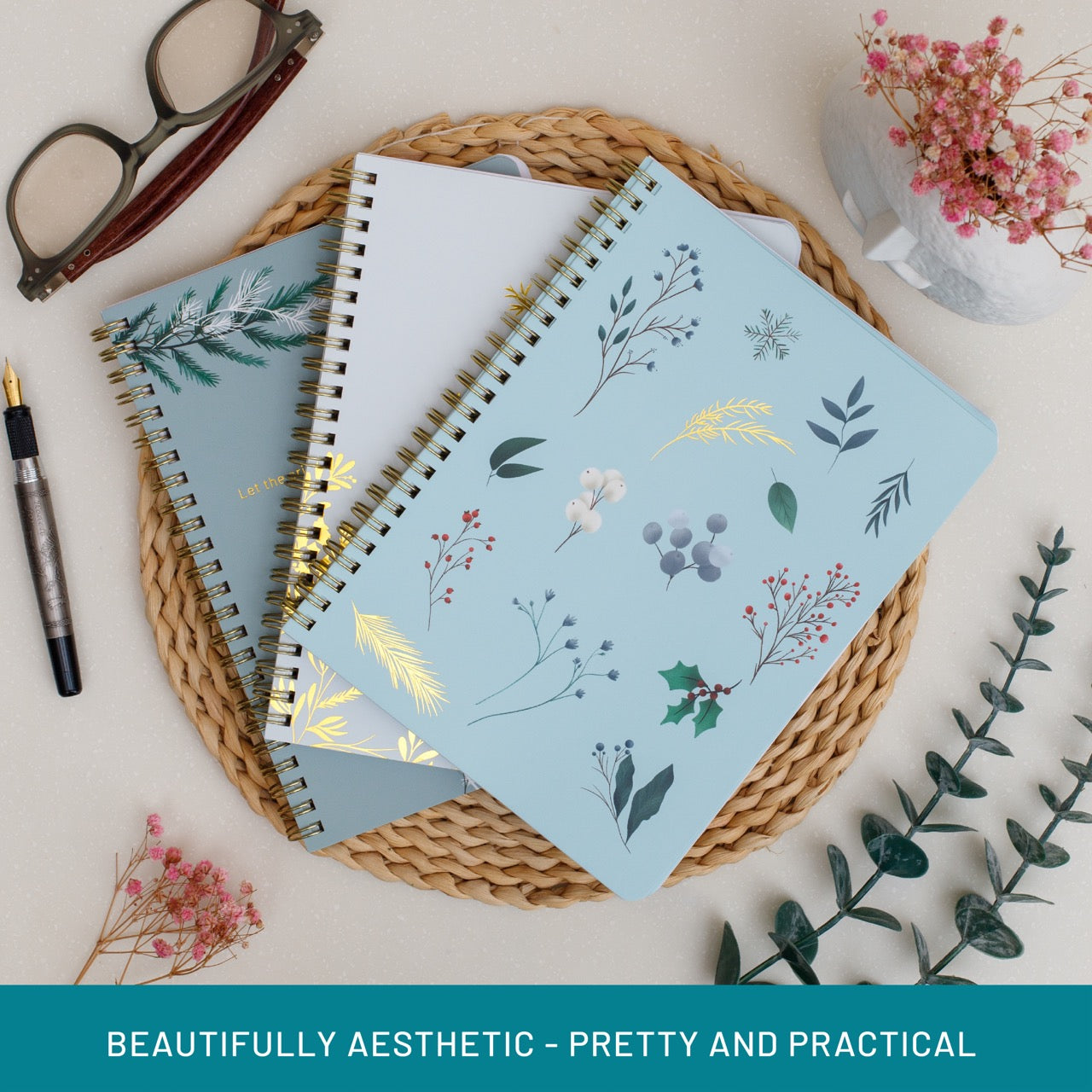 Three notebooks with floral designs on a woven mat with glasses and flowers in the background.