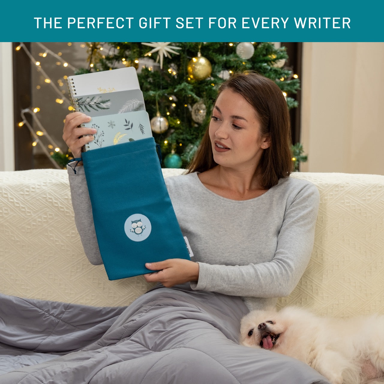 Woman holding a blue bag with 3 notebooks, sitting on a couch with a dog, Christmas tree in the background.