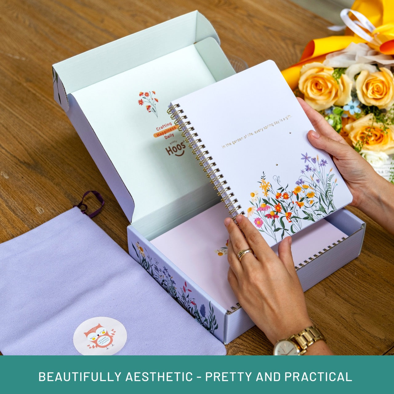 Person holding a notebook with floral design, surrounded by flowers and an open box on a wooden surface.