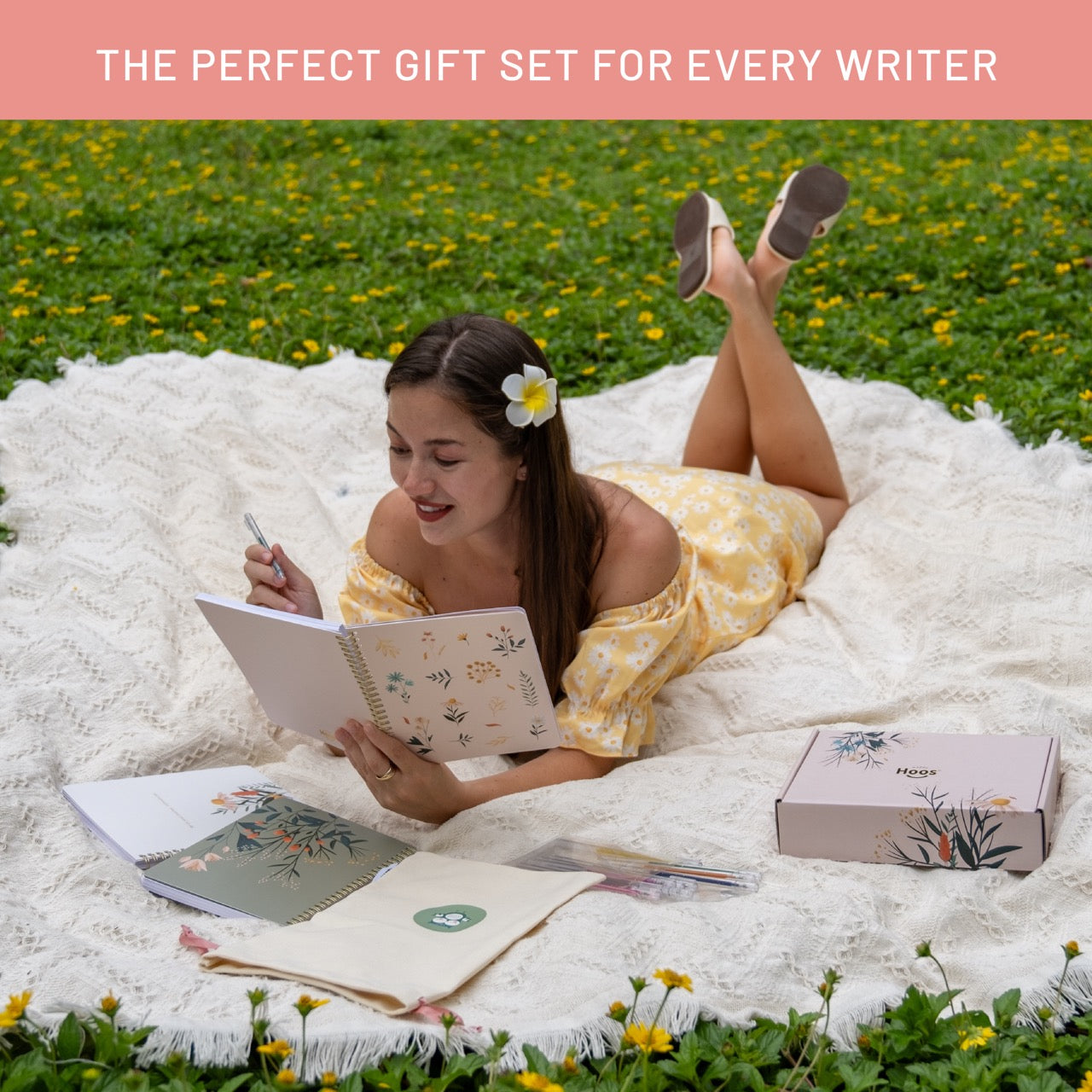 Woman lying on a blanket outdoors, reading a book with stationery items around her.