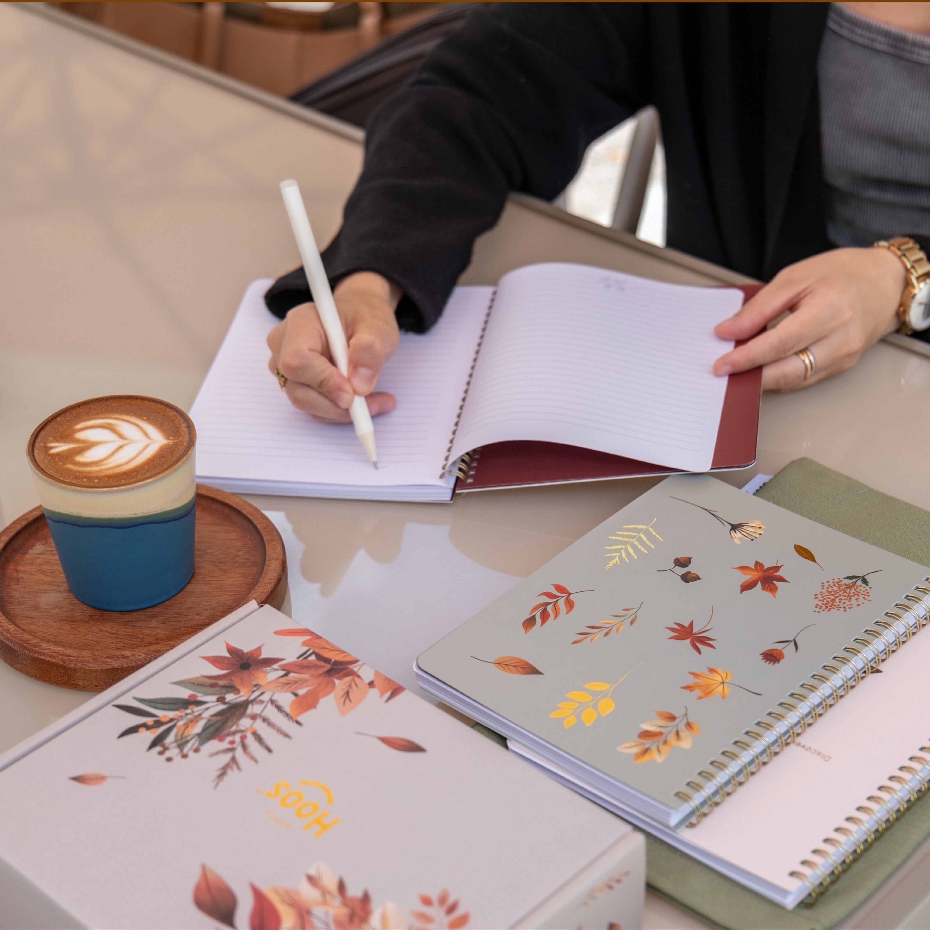 A woman writing in a fall - themed notebook  (Gilded Autumn) with a cup of coffee and two more fall themed notebooks and a fall themed box (Autumn Harvest Gift Set). 