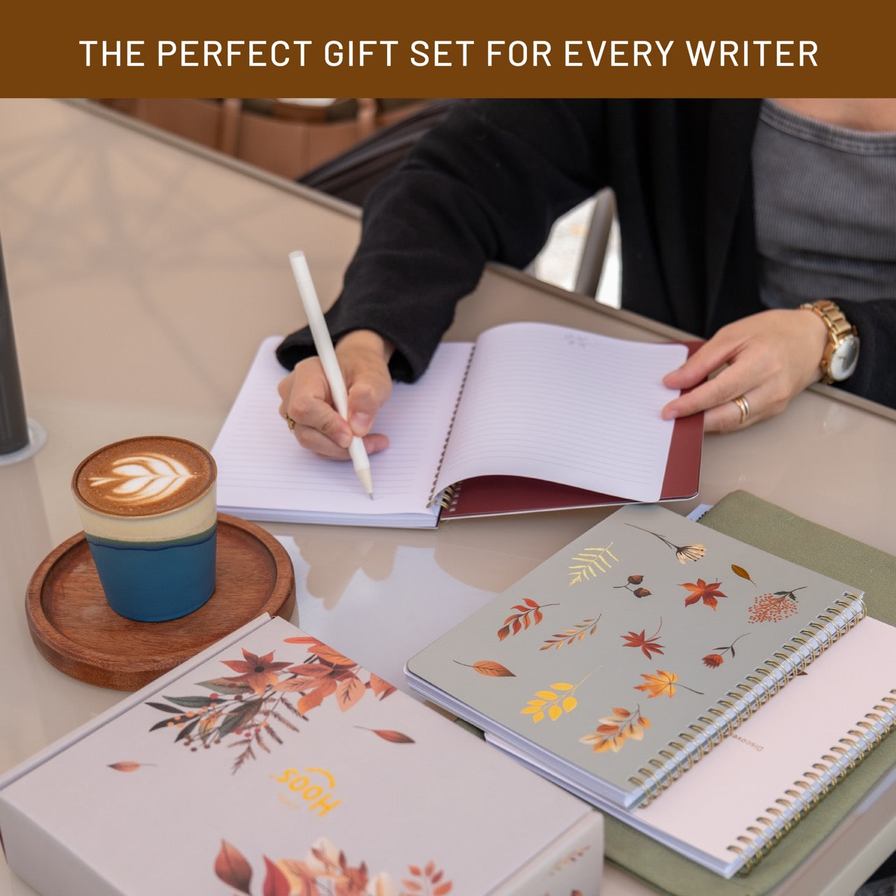 Person writing in a notebook with a cup of coffee and decorative notebooks on a table.