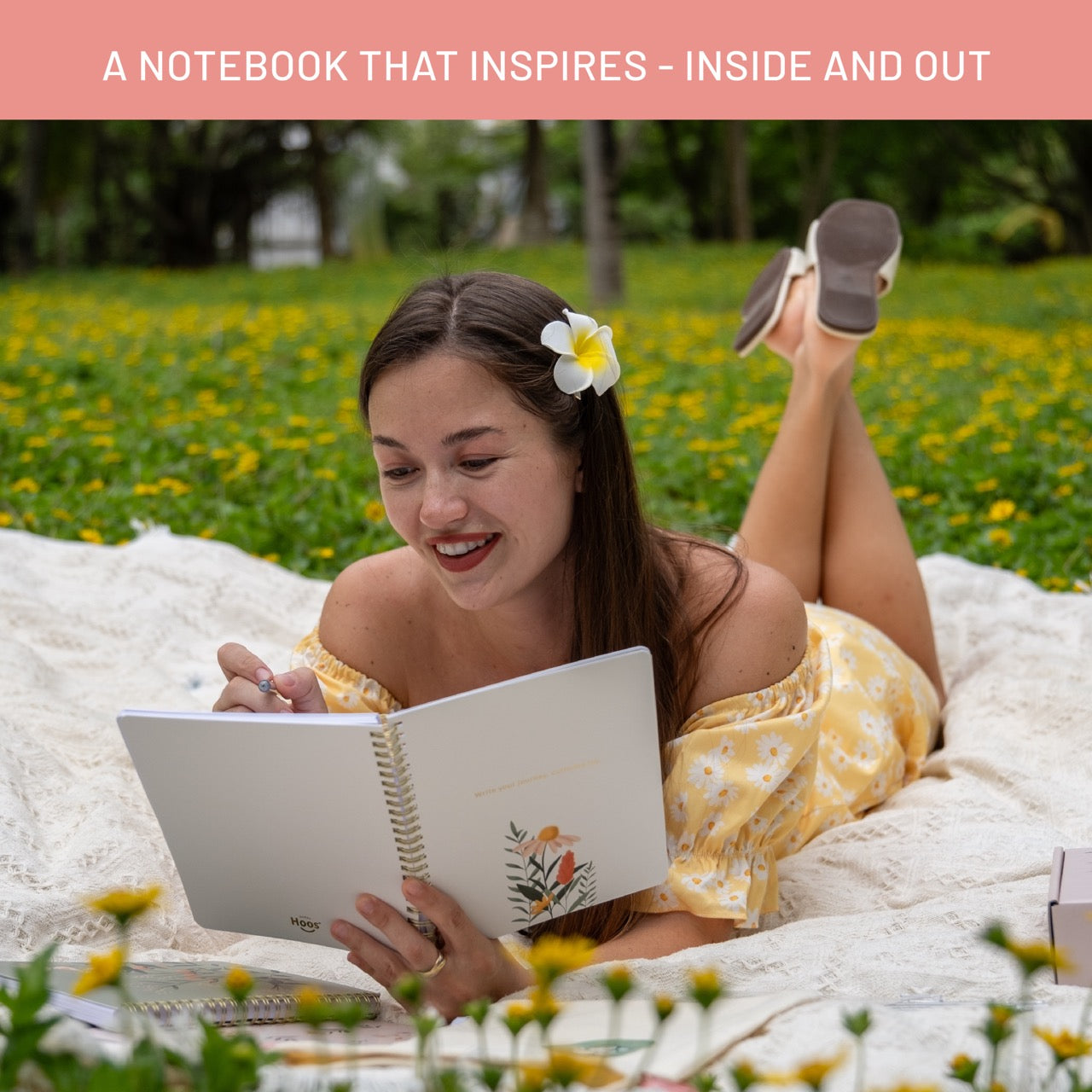 Woman lying on a blanket in a field, reading a notebook with a floral design.