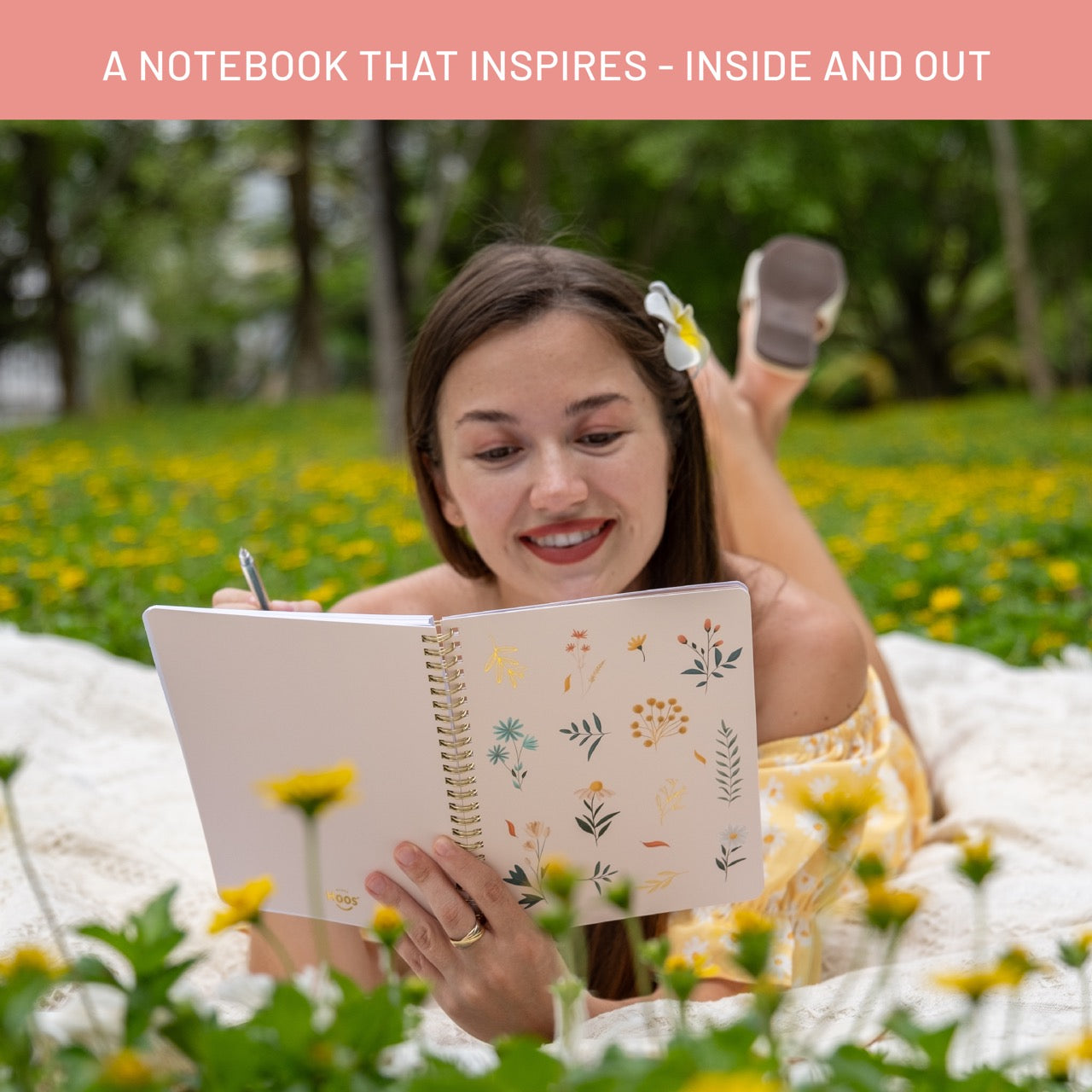Woman lying on a blanket outdoors with a notebook, surrounded by flowers and nature.