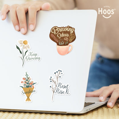 Four mom life stickers on white laptop: 'Brewing Ideas' coffee cup, 'Keep Growing' white daisies, 'Sunshine' flower bouquet, and 'Reset Rise &' blue branches
