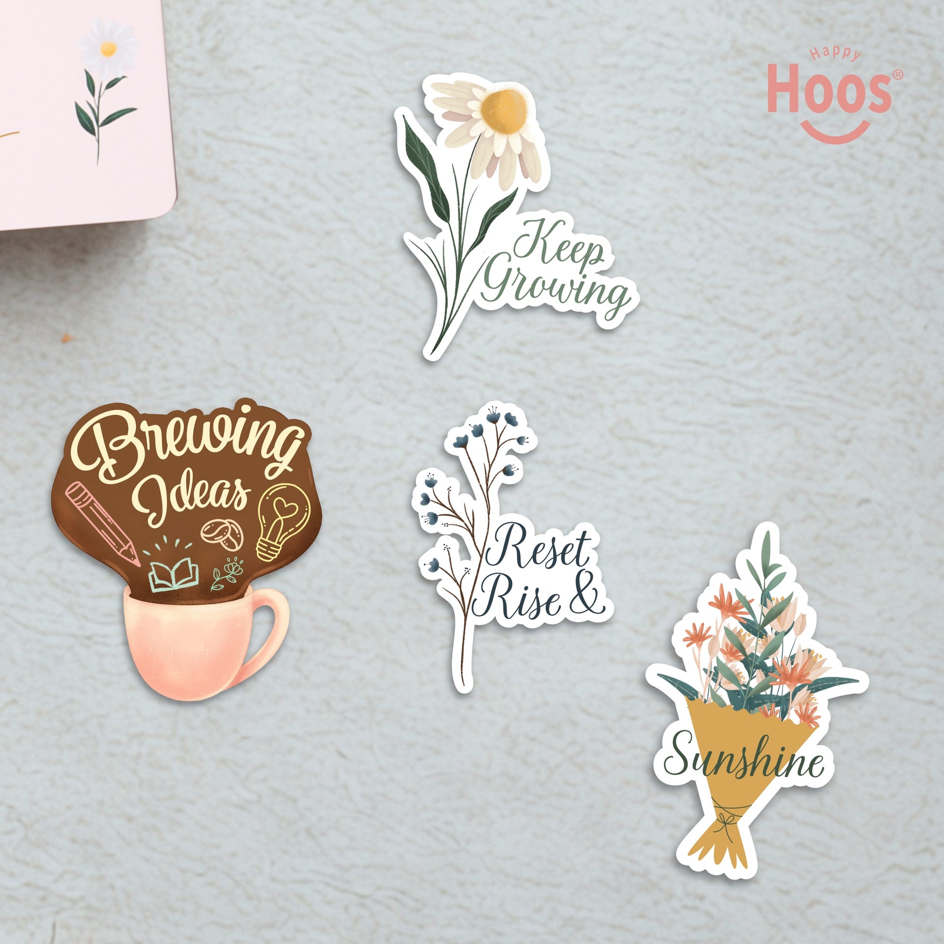 Four motivational stickers on textured surface: 'Keep Growing' white daisies, 'Brewing Ideas' coffee cup, 'Reset Rise &' blue branches, and 'Sunshine' flower bouquet
