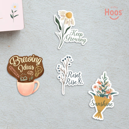 Four motivational stickers on textured surface: 'Keep Growing' white daisies, 'Brewing Ideas' coffee cup, 'Reset Rise &' blue branches, and 'Sunshine' flower bouquet