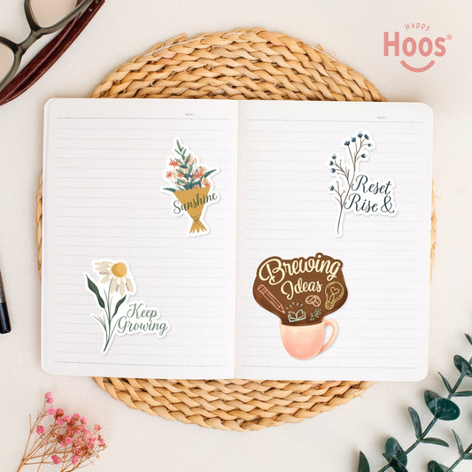 Four stickers in open journal on woven placemat: 'Sunshine' flower bouquet, 'Reset Rise &' blue branches, 'Keep Growing' white daisies, and 'Brewing Ideas' coffee cup