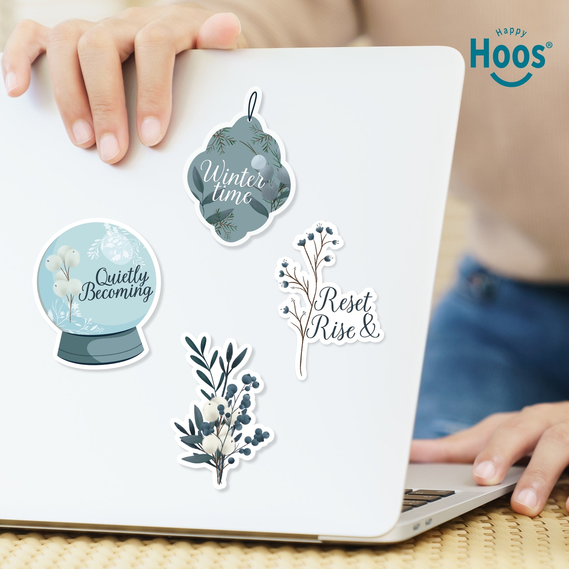 Four winter stickers on white laptop: 'Winter time' ornament, 'Quietly Becoming' snow globe, 'Reset Rise &' branch, and white and blue botanical branch