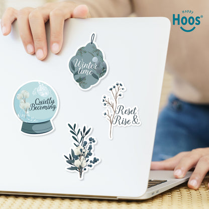 Four winter stickers on white laptop: 'Winter time' ornament, 'Quietly Becoming' snow globe, 'Reset Rise &' branch, and white and blue botanical branch