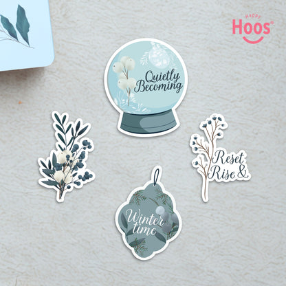 Four winter-themed stickers on textured surface: 'Quietly Becoming' snow globe, white and blue botanical branch, 'Reset Rise &' branch, and 'Winter time' ornament
