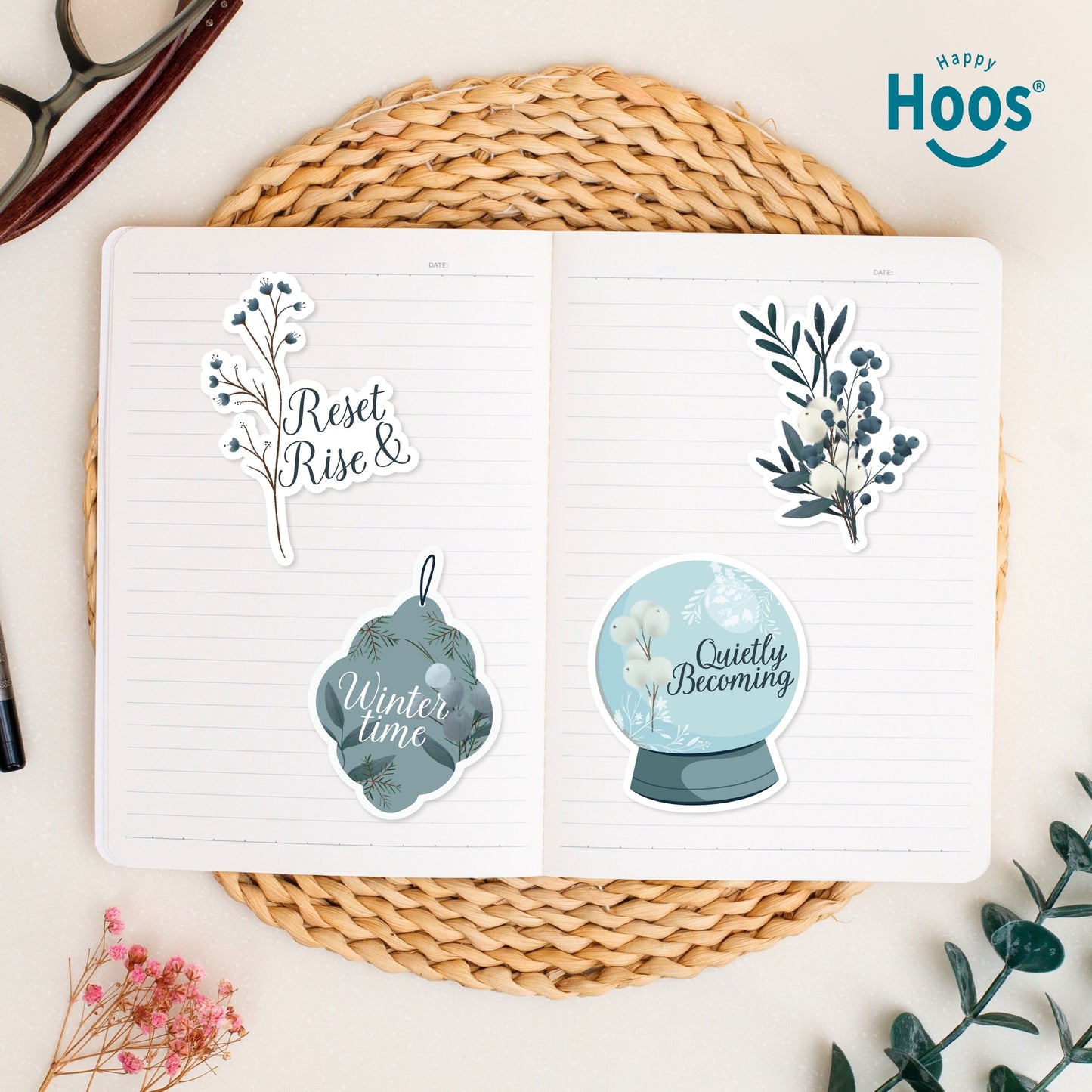 Four stickers in open journal on woven placemat: 'Reset Rise &' branch, white and blue botanical branch, 'Winter time' ornament, and 'Quietly Becoming' snow globe