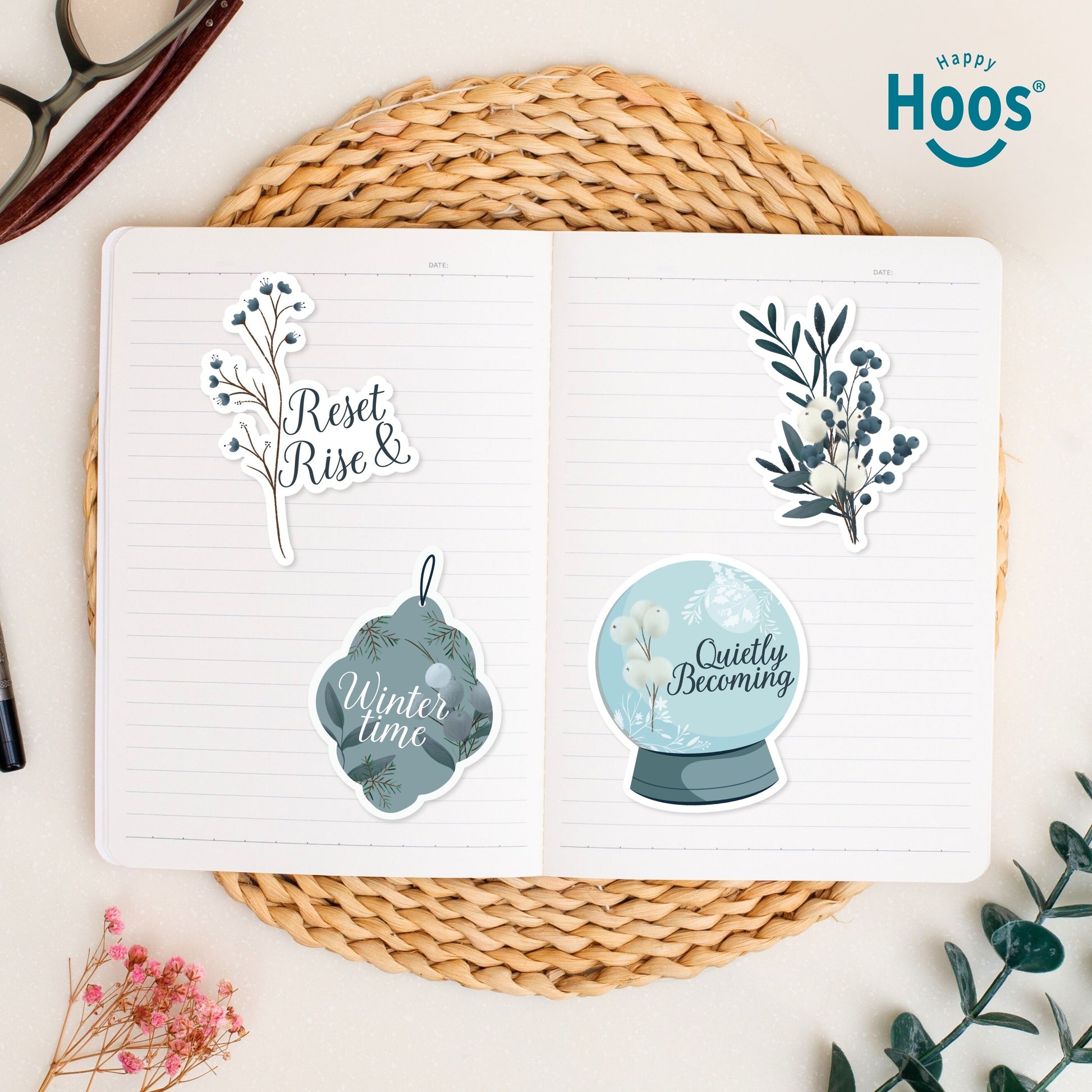Four stickers in open journal on woven placemat: 'Reset Rise &' branch, white and blue botanical branch, 'Winter time' ornament, and 'Quietly Becoming' snow globe