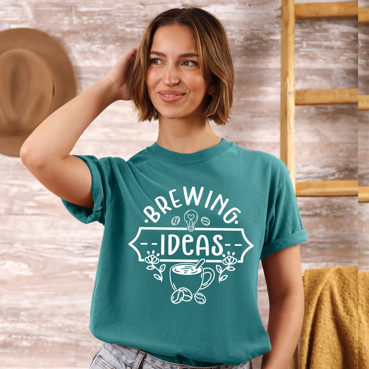 Woman wearing a teal t-shirt with 'Brewing Ideas' design, standing in a rustic setting.
