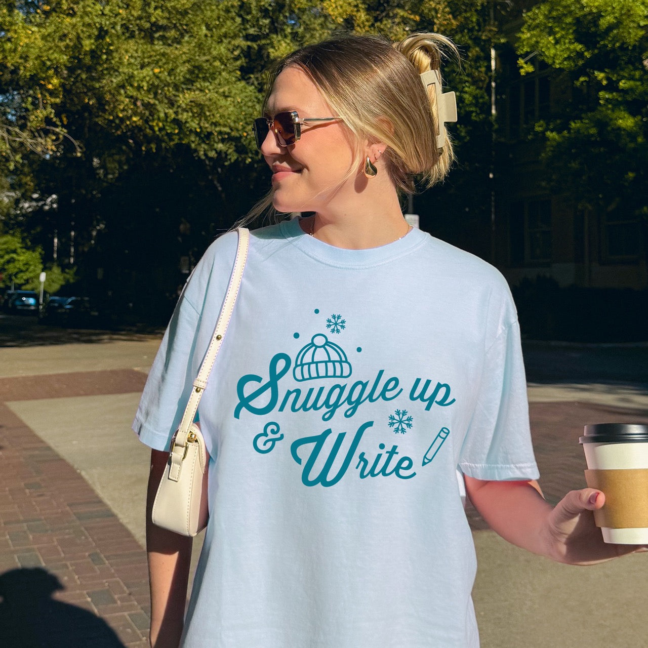 Woman wearing a light blue t-shirt with 'Snuggle up & Write' text, holding a coffee cup.
