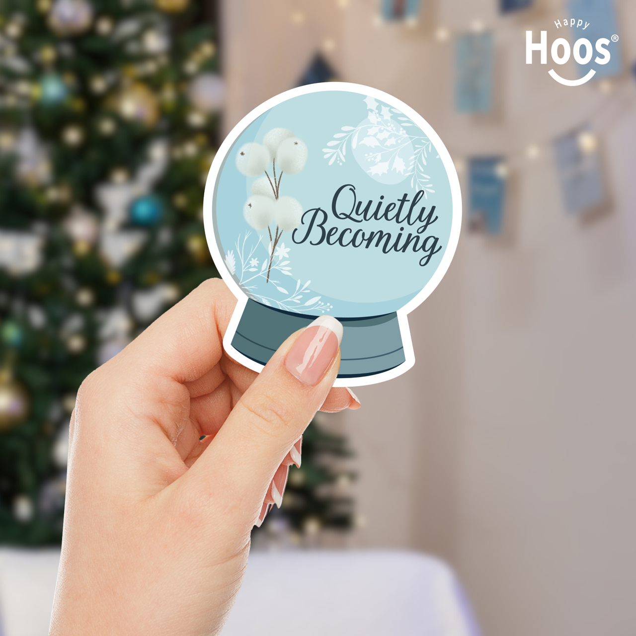 Hand holding a decorative sticker with 'Quietly Becoming' text in front of a Christmas tree.