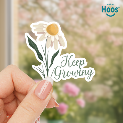 Hand holding a 'Keep Growing' sticker with a daisy design against a blurred floral background.