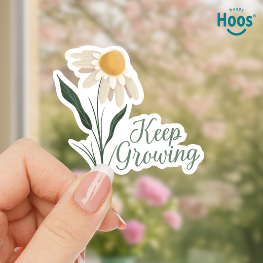 Hand holding a 'Keep Growing' sticker with a daisy design against a blurred floral background.