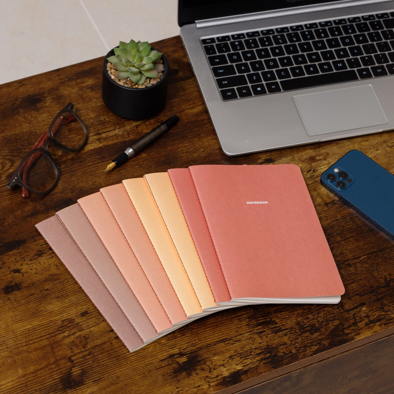 Set of fall colored notebooks on a wooden desk with a laptop, phone, glasses, and pen.