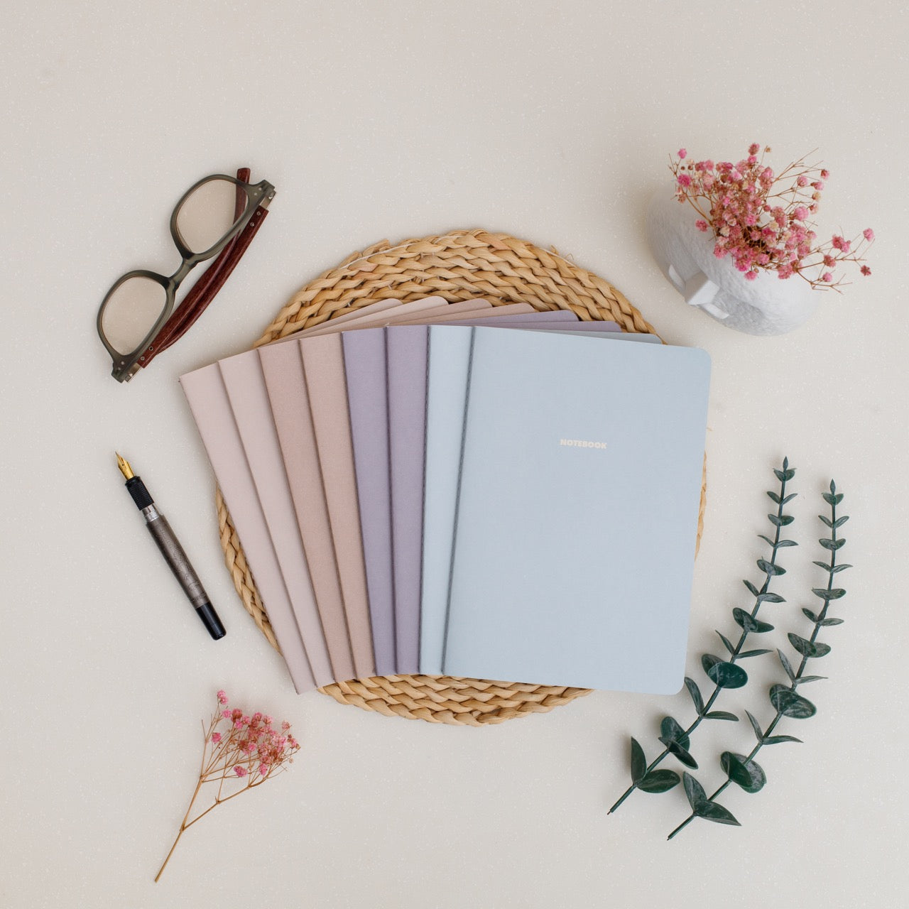 Set of winter pastel-colored notebooks on a woven tray with glasses, pen, and flowers on a light background